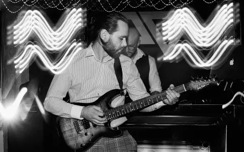 Muzyk gra na gitarze elektrycznej. W tle widać drugiego muzyka i falujące światła, tworzące efekt ekspozycji.