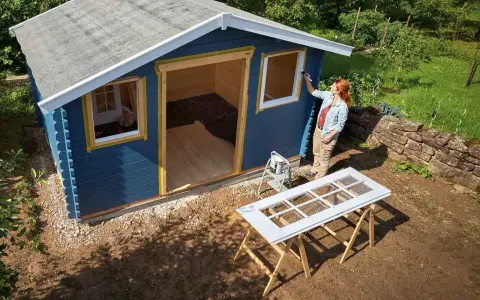Une femme peint les contours des fenêtres d'une cabane de jardin bleue. Le toit de la cabane est en cours de finition.