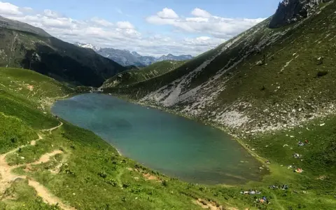 Un sentiero serpeggia tra prati verdi verso il lago Branchino, le cui acque turchesi riflettono le montagne circostanti. Alcune persone si godono un picnic sulle rive.