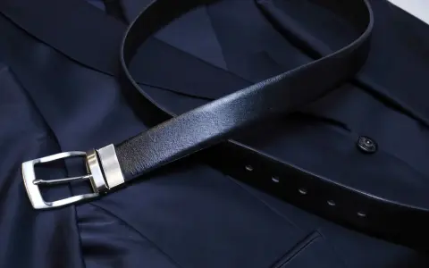 A black leather belt with a silver buckle rests on a dark navy suit jacket. Do you wear a belt with a tuxedo? This image shows a classic accessory.