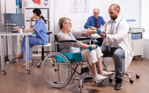 Rehabilitacja po uszkodzeniu móżdżku: pacjentka w wózku wykonuje ćwiczenia z hantlami pod okiem fizjoterapeuty.