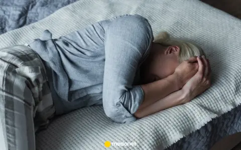 Kobieta w piżamie leży na łóżku, zakrywając twarz dłońmi.