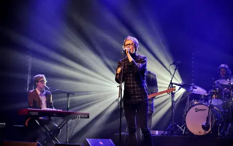 Zespół na scenie, wokalista śpiewa do mikrofonu, klawiszowiec gra na keyboardzie, a perkusista wybija rytm. Wspaniałe tło muzyczne.