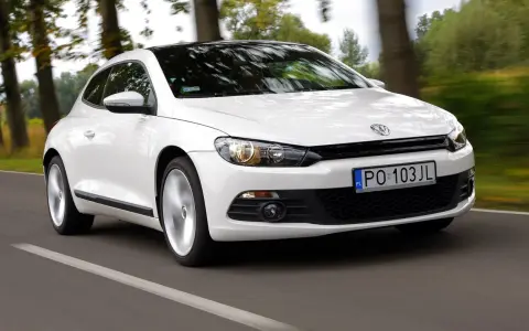 Biały Volkswagen Scirocco pędzi po drodze wśród drzew.