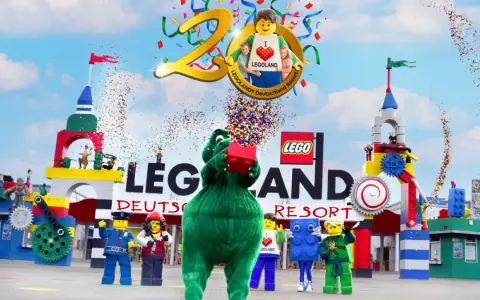 Bunte Konfetti-Explosion bei der Legoland Parade! Ein grüner Drache hält einen roten Lego-Stein hoch, umgeben von Figuren und bunten Türmen.