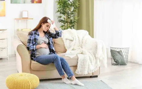 Donna incinta seduta sul divano con un fazzoletto sulla fronte, che soffre di raffreddore in gravidanza.