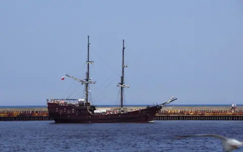 Drewniany statek z masztami i żaglami, przypominający piracki, pływa po wodzie. To tramwaj wodny Darłowo, pełen turystów.