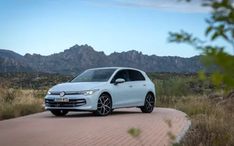 Un coche compacto Volkswagen Golf blanco circula por una carretera de adoquines con montañas rocosas al fondo.