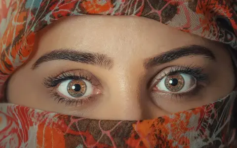 Ojos color miel que miran con curiosidad, enmarcados por un velo con estampado naranja y marrón.