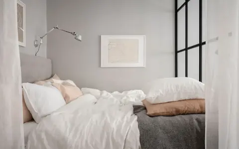 En inbjudande säng med vita och ljusrosa kuddar, en grå pläd och en konstnärlig tavla på väggen. En liten alkov som känns ombonad.