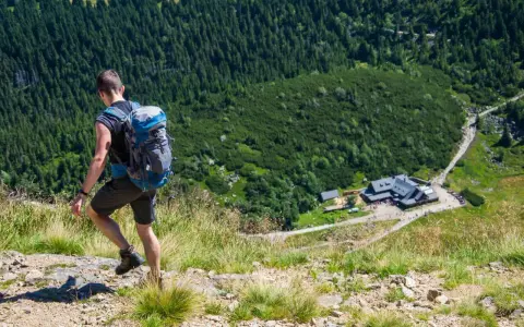 Turysta z plecakiem schodzi po kamienistej ścieżce w Karkonoszach. W dole widoczny schronisko, jedna z wielu atrakcji górskich.