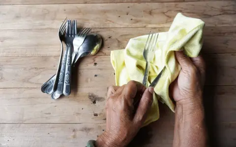 Mains polissant des couverts en argent avec un chiffon jaune. Un tas de fourchettes et cuillères est à gauche. Apprenez comment nettoyer l'argenterie.