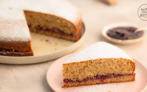 Eine köstliche, schnelle Buchweizentorte mit Marmeladenfüllung und Puderzucker bestäubt. Ein Stück auf einem rosa Teller, der Rest auf einem größeren Teller.