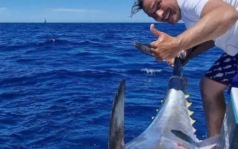 Uomo felice mostra il suo **biggest tuna** appena pescato in mare aperto, con una barca e una vela all'orizzonte.