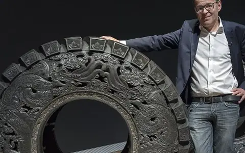 Wim Delvoye pose à côté d'une roue ornée de dragons, une œuvre d'art monumentale.