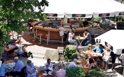 Gäste sitzen an Tischen und an der Theke eines Biergartens unter Sonnenschirmen.