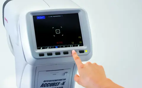 Dłoń naciska przycisk na auto refraktometrze, pokazując ekran z danymi badania wzroku. Pozwala to zrozumieć, jak odczytać wydruk z komputerowego badania wzroku.