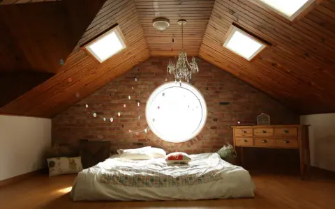 Camera puntata verso un letto in mansarda, con il soffitto in legno e la sua evidente pendenza tetto. Finestre sul tetto e un oblò circolare illuminano la stanza.