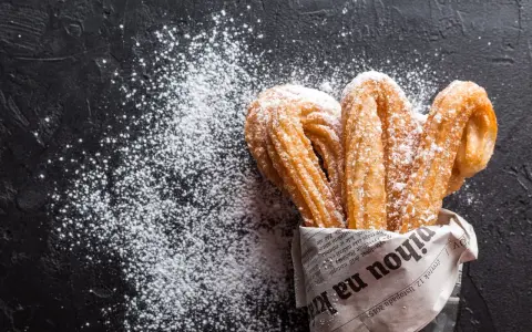 Churros z czekoladą kontra pączki z lukrem? Te złociste churros, posypane cukrem pudrem, kuszą na czarnym tle.