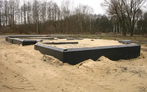 Fundament domu w piasku, widoczne czarne membrany izolacyjne i drewniane elementy szalunku. Warstwy gruntu przygotowane do dalszych prac budowlanych.