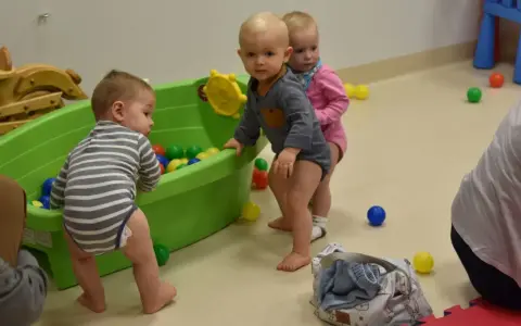 Trzy maluchy, w tym 6 miesięczne dziecko w chodziku, bawią się kolorowymi piłeczkami w zielonej wanience.