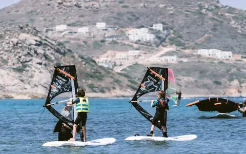 Kinder lernen naxos surfen. Zwei junge Windsurfer üben auf dem Meer, im Hintergrund weiße Häuser am Hang.