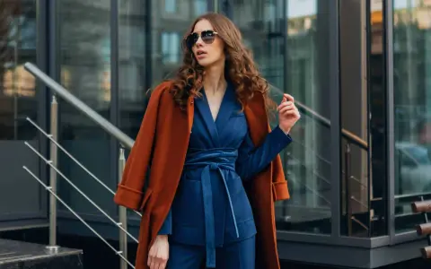 Une femme porte un gilet avec une robe bleu marine, un manteau orange sur les épaules, et des lunettes de soleil.