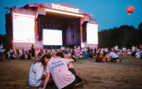 Para siedząca na trawie przed sceną festiwalową z napisem "bittersweet". W tle tłum ludzi i wielkie ekrany.