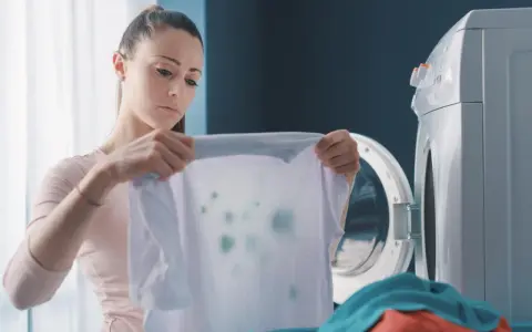 Zmartwiona kobieta trzyma białą koszulkę z zielonymi plamami, która wygląda jak zafarbowane ubrania. Obok stoi otwarta pralka.