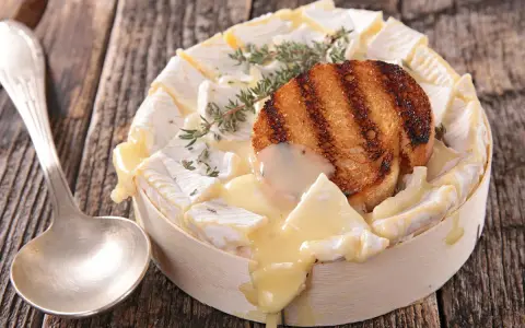 Un camembert coulant, plat typique normandie, garni d'une tranche de pain grillé et de thym, prêt à être dégusté avec une cuillère.