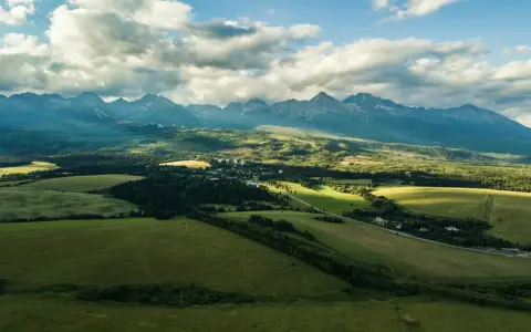 Malowniczy krajobraz z zielonymi polami i lasami, w oddali majestatyczne słowacja góry pod błękitnym niebem z chmurami.