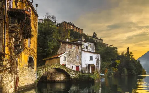 Malerische Häuser am Wasser in Nesso am Comer See, mit einer alten Steinbrücke und goldenem Abendlicht.