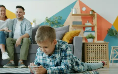 Chłopiec gra na telefonie, podczas gdy rodzice siedzą na kanapie. Ciekawe, od ilu lat jest WhatsApp i czy oni też go używają?