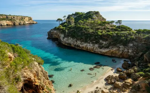 Malownicza zatoczka z turkusową wodą i piaszczystą plażą, otoczona klifami i zielenią. Idealne miejsce na relaks, jedna z pięknych palma de mallorca atrakcje.