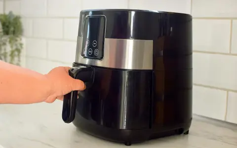 En hand drar ut korgen på en svart airfryer. Den här köksapparaten är lika praktisk som en diskmaskin för snabb matlagning.