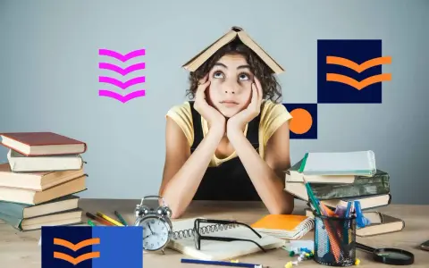 Zmęczona studentka z książką na głowie, otoczona stosami książek i materiałami do nauki. Tak wyglądają studia niestacjonarne.