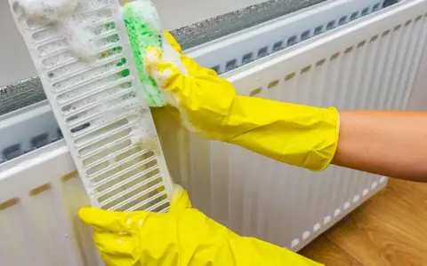 Manos con guantes amarillos limpian la rejilla de un radiador con una esponja y espuma.