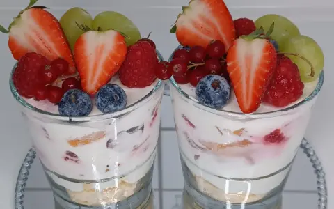 Zwei Gläser mit einem cremigen Dessert, garniert mit frischen Erdbeeren, Himbeeren, Blaubeeren, Johannisbeeren und Trauben. Ein köstliches Dessert im Glas.