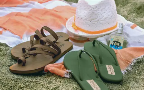 Brown sandals vs. green flip flops on a beach towel, with a hat and body mist. A perfect beach day awaits!