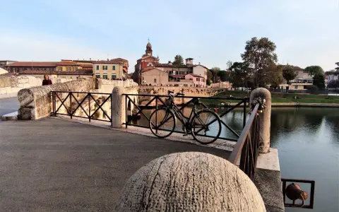 Ponte sul fiume a Borgo San Giuliano, con una bicicletta appoggiata alla ringhiera e palazzi sullo sfondo.
