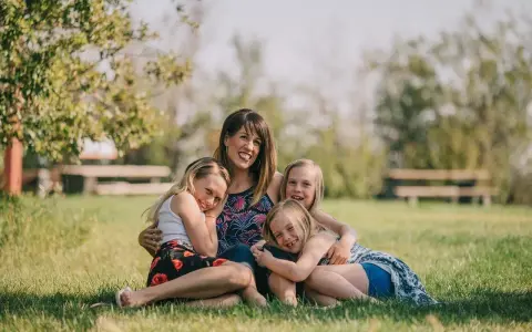 Mama z córkami na trawie, uśmiechnięte i przytulone. Widać, że czują motyle w brzuchu od tej miłości.