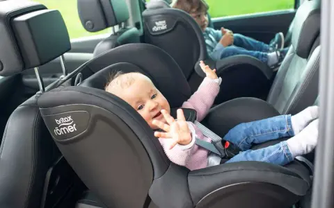 Uśmiechnięte niemowlę w foteliku Britax Römer, obok starsze dziecko. Od kiedy dziecko może jeździć z przodu? To pytanie nurtuje wielu rodziców.
