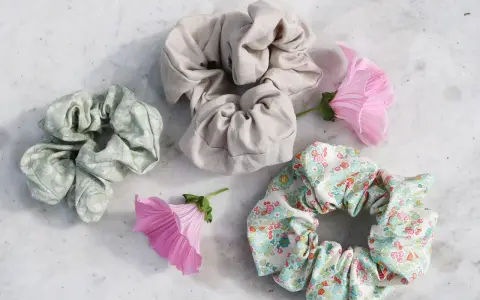 Tre virka scrunchies i olika mönster och färger, omgivna av rosa blommor på en marmorerad yta.