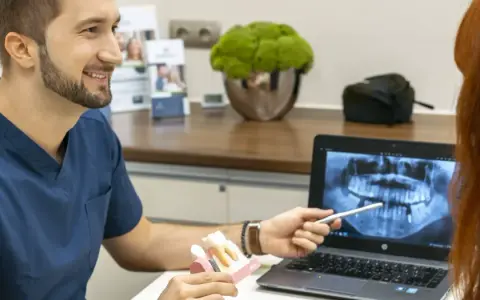 Dentysta omawia z pacjentką plan leczenia, pokazując na ekranie zdjęcie rentgenowskie szczęki i model zębów, wyjaśniając znaczenie czyszczenia dziąseł.