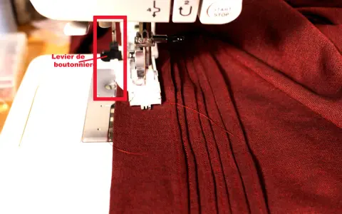 Levier de boutonnière sur machine à coudre, prêt pour réaliser une boutonnière parfaite sur un tissu bordeaux plissé.