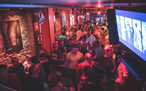 Un locale affollato a Barcellona by night, con uomini che socializzano e bevono. L'atmosfera è vibrante, illuminata da luci rosse e blu.