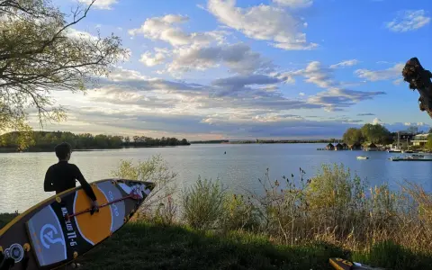 Ein junger Mann mit SUP-Board am Ufer des Alperstedter Sees. Der Himmel ist blau mit weißen Wolken, die Sonne scheint.