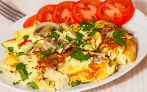 Pyszny omlet jajeczny z pieczarkami i natką pietruszki, podany z plastrami pomidora. Idealny na śniadanie.