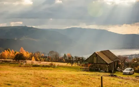 Jesienne słońce przebija się przez chmury nad hrobcza łąka. Stara chata, drzewa w złotych barwach i jezioro w oddali.