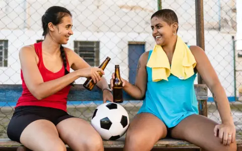 Zwei Frauen mit Fußball und Bierflaschen stoßen an. Nach dem Sport ist vor dem Genuss: sport und alkohol.